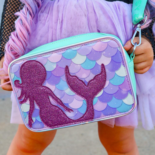 A close up of a girl wearing a green crossbody bag with an interchangeable cover with a purple glitter mermaid on a purple and green scales background.