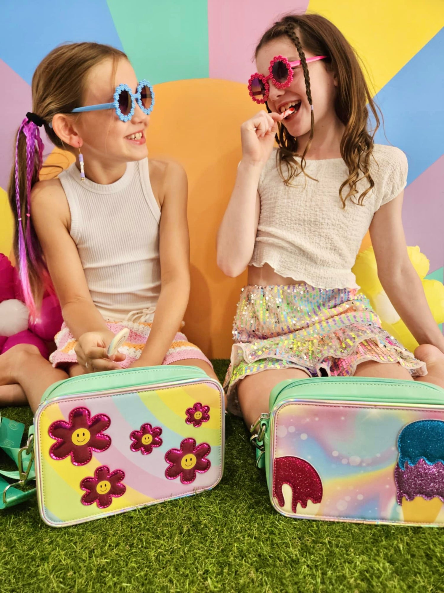 Two girls sitting in front of a colorful background, licking lollipops, each with a kids mint crossbody bag. The girl on the left has the Flower Fields cover, and the girl on the right has the Sundae Fundae ice cream cover.