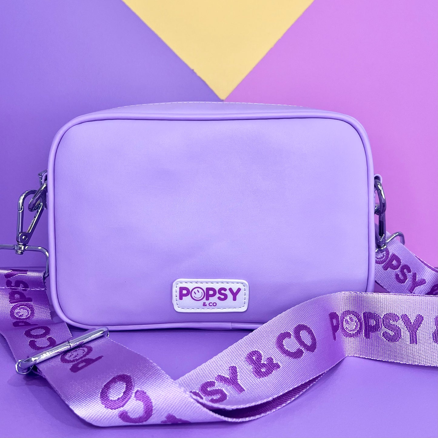 Purple kids crossbody bag. Has magnetic clips on front to attach a Popsy interchangeable cover and logo on the back. Has an adjustable strap.