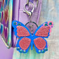 Butterfly keychain for kids. It has a purple metallic butterfly with pink glitter patches and blue gems.