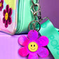 Pink flower bag charm with a yellow smiley in the middle. It is clipped onto a Popsy & Co crossbody bag.